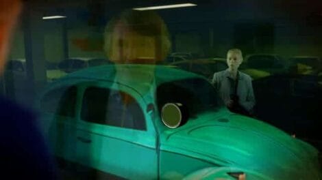 Image: A man's reflection is visible on the glossy surface of a turquoise vintage car in a Hollywood parking garage, with another man standing in the background. -- D.C. Douglas