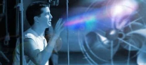 Image: An engineer in a tank top uses a touchscreen interface with holographic projections of technology and data surrounding him in a futuristic blue-toned laboratory setting as "My Art Hits The Fan" illuminates the background -- D.C. Douglas
