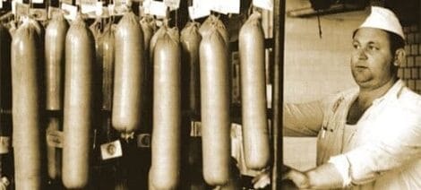 Image: A black and white photo of a butcher in a cap standing next to several large hanging sausages in a shop, acting like he's in an old-time movie. -- D.C. Douglas