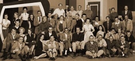 Image: A vintage black and white photo of a large group of Estelle Harman Alumni, approximately thirty, posed together in several rows, smiling, dressed in mid-20th century attire, in what appears -- D.C. Douglas