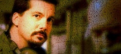 Close-up of a man with facial hair looking towards the camera in a dimly lit environment.