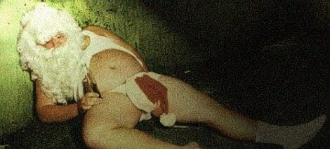 Image: A person dressed as Santa Claus is slumped against a wall, appearing distressed or unconscious, holding a bottle in a dimly lit setting, potentially egg nog intolerant. -- D.C. Douglas