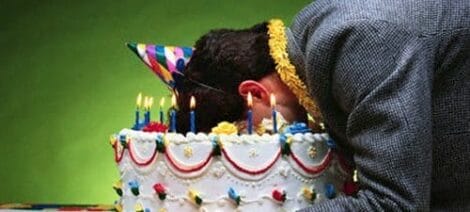 Image: A man wearing a party hat presses his face comically into a large birthday cake adorned with lit candles and colorful decorations, humorously acting his age. -- D.C. Douglas