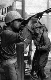 Image: Black and white photo of two soldiers in helmets; one aiming upward with a rifle and the other watching intensely, designed as a scene from a video game, standing in a battle-scarred environment. -- D.C. Douglas