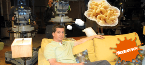 Image: A man lounges on a yellow sofa on a TV set with cameras in the background. He is imagining a cloud of macaroni above him, accompanied by the Nickelodeon logo with an orange -- D.C. Douglas