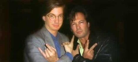 Image: Two men posing together at night, one wearing a gray blazer and glasses, the other in a black leather jacket designed by Eric Cotsen, both using their hands to make a symbolic gesture. -- D.C. Douglas