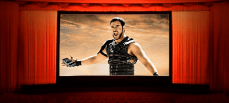 Image: A man dressed in gladiator attire holds a remote control, expressing a shout of victory or challenge, displayed on a cinema screen with an entertainment-themed red curtain frame. -- D.C. Douglas