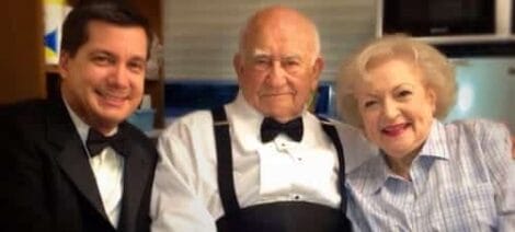 Image: Three elderly individuals smiling for the camera, seated at a table. The man in the middle wears a tuxedo with a bow tie, flanked by another man in a suit and an elderly woman -- D.C. Douglas