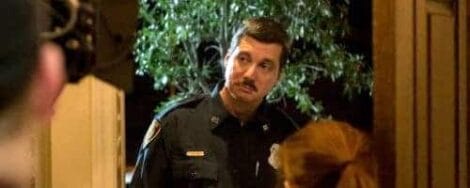 Image: A police officer in uniform speaking to a person, partially seen in the foreground, in a dimly lit indoor setting with a plant and Hollywood poster visible in the background. -- D.C. Douglas