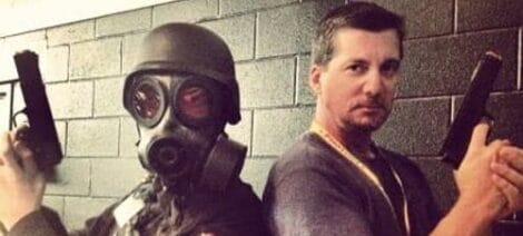 Image: Two men posing against a gray brick wall at Florida SuperCon; one wears a gas mask and holds a pistol, while the other, wearing a casual shirt and necklace, also holds a pistol. -- D.C. Douglas