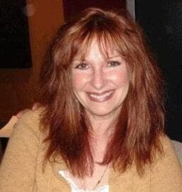 Image: A smiling woman with shoulder-length reddish-brown hair, wearing a mustard-colored sweater, poses for the camera in a warmly lit indoor setting while holding a video game controller. -- D.C. Douglas