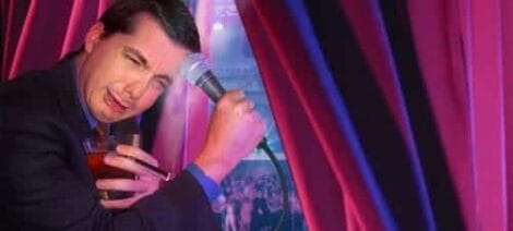 Image: Man peeking through red stage curtains, holding a microphone and a cocktail, with a crowd visible in the background, looking amused and curious. -- D.C. Douglas