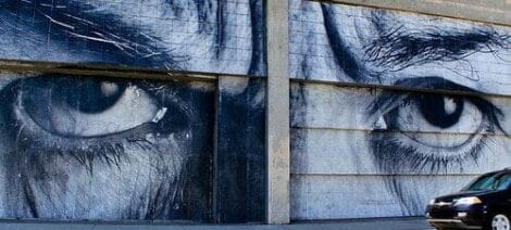 Image: A striking mural of a pair of intense, realistic eyes painted on a segmented concrete wall, showcasing the artist's creativity, with a car passing by in the foreground. -- D.C. Douglas