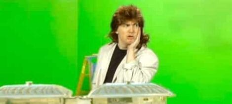 Image: A man with a shaggy hairstyle wearing a white lab coat stands in front of a green screen, looking surprised with a hand near his face. There are metal containers in the foreground, used as -- D.C. Douglas