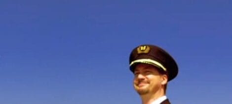 Image: A pilot smiling in uniform, set against a clear blue sky. His cap features a golden emblem resembling the Titanic, and he is looking slightly to his right. -- D.C. Douglas