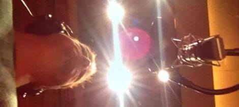 Image: View from behind of a resident actor wearing headphones facing a microphone, with a bright studio light creating a lens flare effect. -- D.C. Douglas