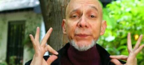 Image: An older man with a shaved head and a maroon turtleneck makes a playful face and gesture with his hands near his cheeks, standing outdoors with trees and a building in the background. He is -- D.C. Douglas