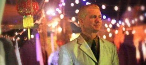 Image: A man in a white suit stands partially out of focus at a night event, illuminated by colorful blurred lights hanging in the background of a dystopian fiction setting. -- D.C. Douglas