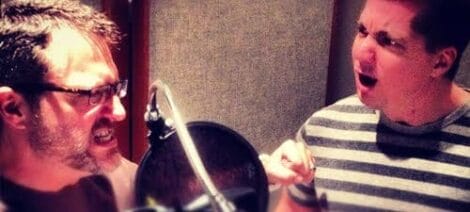 Image: Two men excitedly recording a voiceover in a studio, one is shouting into a microphone while the other listens wearing headphones, displaying animated expressions in a soundproof room. -- D.C. Douglas