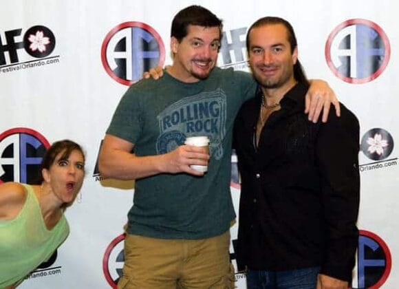 Image: Two men pose smiling for a photo at an event, one holding a drink. A woman playfully photobombs them on the left side. They stand in front of a banner emblazoned -- D.C. Douglas