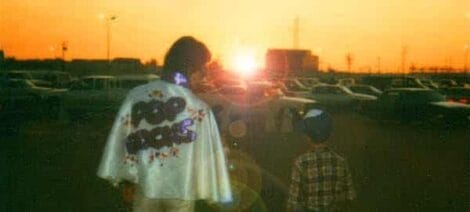Image: A person and a child stand in a parking lot, gazing towards a brilliant sunset. The person wears a jacket with "Pop Rocks" on the back. -- D.C. Douglas