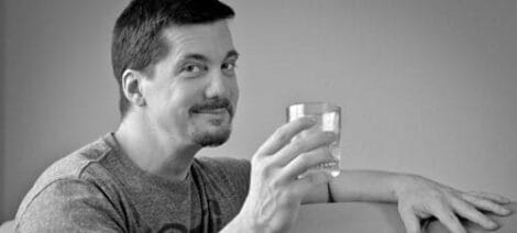Image: A man with a slight smile holding a glass of water while sitting at a table, depicted in a black and white photo, exuding a Wicked Mojo. -- D.C. Douglas