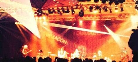 Image: A lively rock concert scene at Youmacon with a band performing on stage, highlighted by bright stage lights and a large crowd in silhouette. -- D.C. Douglas