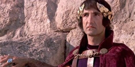 Image: A king in a medieval costume, featuring a purple robe and golden crown, stands against a rocky background, looking intently to the side. -- D.C. Douglas
