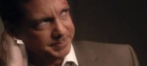 Image: A close-up image of a tan man squinting suspiciously, wearing a suit and tie, with a dimly lit background suggesting an indoor setting. He appears to be in a thoughtful or questioning -- D.C. Douglas