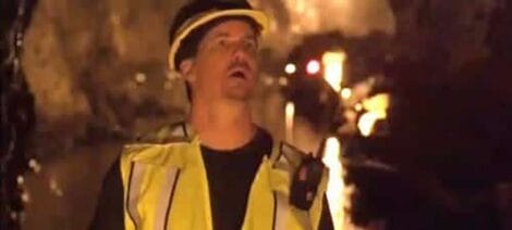 Image: A man in a yellow reflective safety vest and cap looks upwards with an expression of concern or surprise, illuminated by ambient firelight where a Sharknado has just landed in the background. -- D.C. Douglas