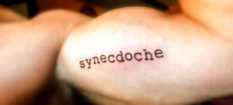 Image: Close-up of a tattoo on a person's upper arm, featuring the literary device "synecdoche" in black, typewriter-style font. -- D.C. Douglas