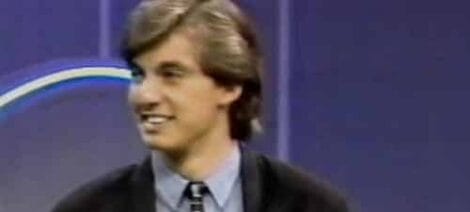 Image: A young man with shoulder-length brown hair and wearing a dark suit with a tie appears on the vintage television show "My Career Now" with a blurred colorful logo in the background. -- D.C. Douglas