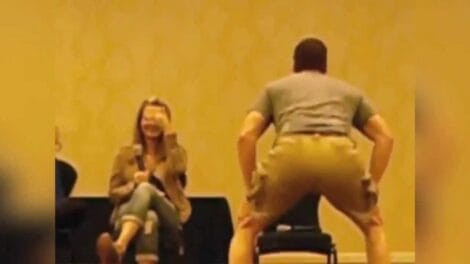 Image: A man wearing gray clothes is standing with his back to the camera, facing a seated woman who is laughing at his twerking. They are in a room with beige walls and appear to be at -- D.C. Douglas