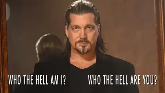 Image: A man with shoulder-length hair and a goatee stands in a black shirt and looks intently towards the camera, with a reflective text overlay on a mirror behind him reading "D.C. Douglas who -- D.C. Douglas