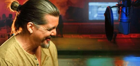 Image: A man with a ponytail, smiling while engaging in a PR event and speaking into a microphone in a radio studio, with colorful blurred lights in the background. -- D.C. Douglas