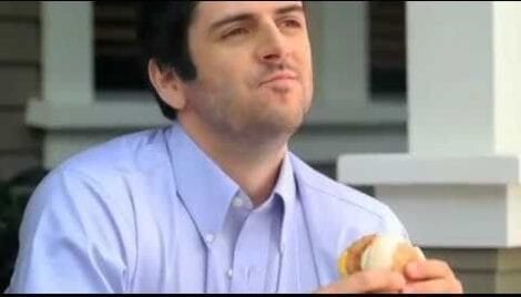 A man in a blue shirt holding a hamburger while sitting outdoors with a thoughtful expression.