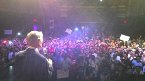 Crowd of people at a Ron Paul speaking event with signs and lights in the background.