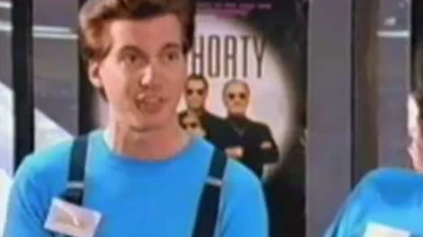 A man wearing a blue shirt and suspenders speaking in front of a poster featuring two men in sunglasses.