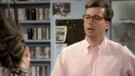 Man wearing a pink shirt and glasses, appearing surprised in a room with shelves of media.