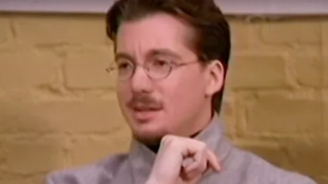 Man wearing glasses and a mustache, dressed in a gray shirt, seated and looking thoughtful.