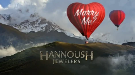 Red hot air balloon shaped like a heart with 'Marry Me!' text floating above a mountainous landscape.
