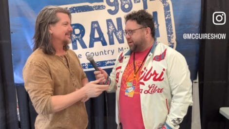 Two men talking with a microphone at an event, one wearing a light jacket and the other in a casual shirt.
