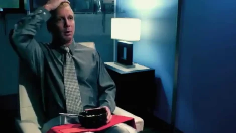 A man in a gray shirt and tie sitting in a chair, holding a bowl and looking surprised.
