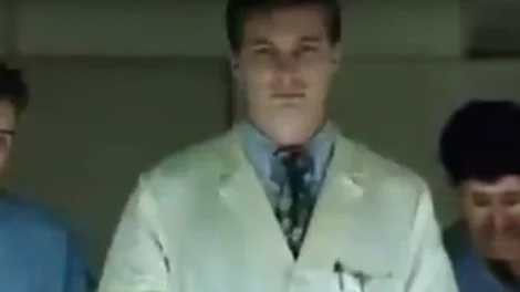 A man wearing a white lab coat and a tie, standing in a neutral pose.