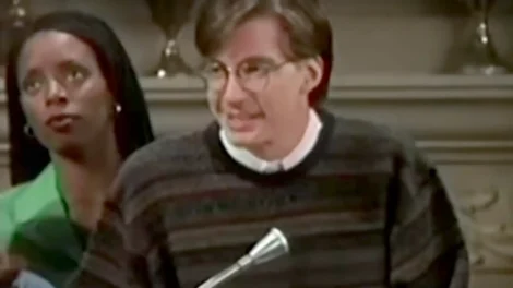 A man with glasses and a striped sweater speaking at a podium during a presentation.