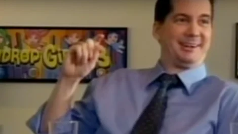 Man wearing a dress shirt and tie, smiling and gesturing with his hand in an indoor setting.