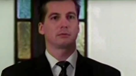Man wearing a black suit and tie standing in front of stained glass windows.