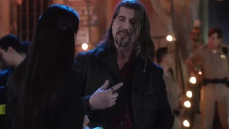 Man with long hair and a beard wearing a dark coat, engaged in conversation.