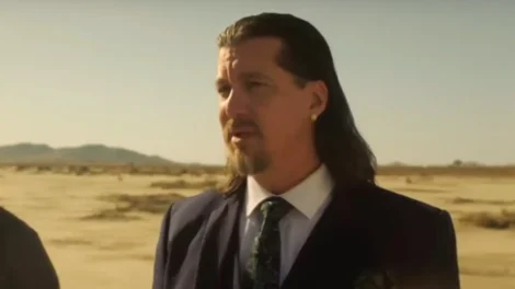 A man with long hair wearing a suit and tie, standing outdoors in a dry landscape.
