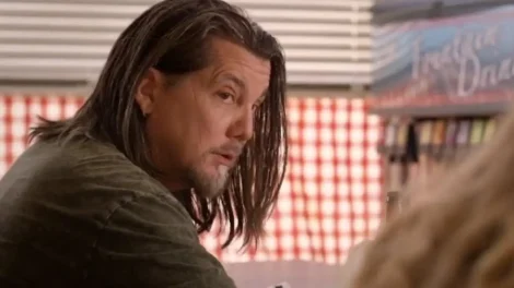 Man with long dark hair sitting at a table, looking towards the camera with a serious expression.